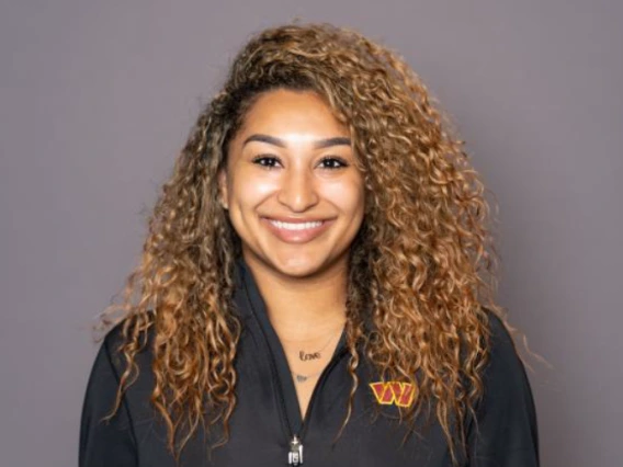 Portrait of a smiling woman with long, curly light brown hair, wearing a black zip-up jacket with a red and yellow embroidered logo on the left side. She stands against a neutral gray background.