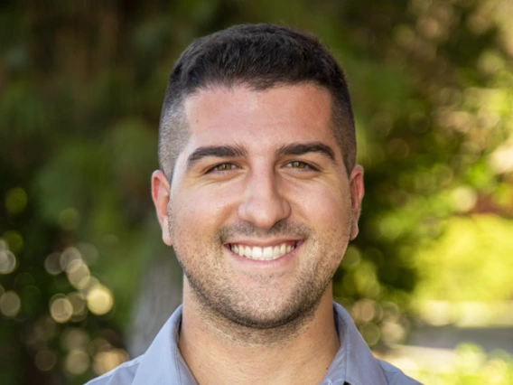 Portrait of a smiling man with short dark hair, wearing a light gray button-up shirt. He stands outdoors with a blurred background of green trees and sunlight filtering through the leaves.