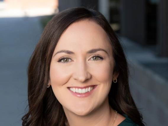 Professional headshot of a woman with long, wavy brown hair, wearing a dark green blouse, gold earrings, and a delicate gold necklace. She is smiling and standing outdoors with a blurred modern building in the background.