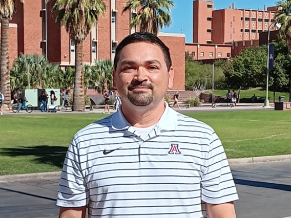 Portrait of a man with short dark hair and a goatee, wearing a white and navy striped polo shirt with a University of Arizona logo. He is standing outdoors on a university campus with palm trees, red brick buildings, and students walking in the background under a bright blue sky.
