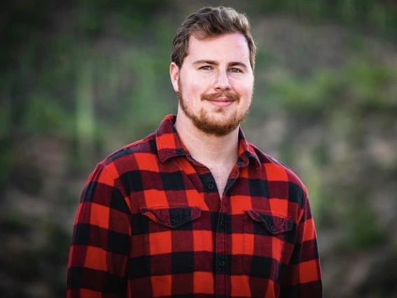 A young man with short brown hair and a trimmed beard stands outdoors in front of a blurred, green and rocky background. He is wearing a red and black plaid flannel shirt and smiling softly at the camera.