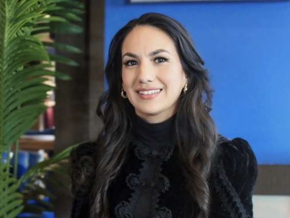 Smiling woman with long dark wavy hair, wearing a black textured high-neck top, seated indoors with a vibrant blue wall and green plant in the background.