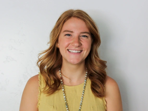 Mikay Wright smiling in front of a light gray wall, wearing a yellow sleeveless top and a beaded necklace