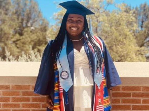 Smiling woman in graduation attire wearing a blue cap and gown, white dress, and multiple colorful stoles, standing in front of a brick wall with trees in the background.