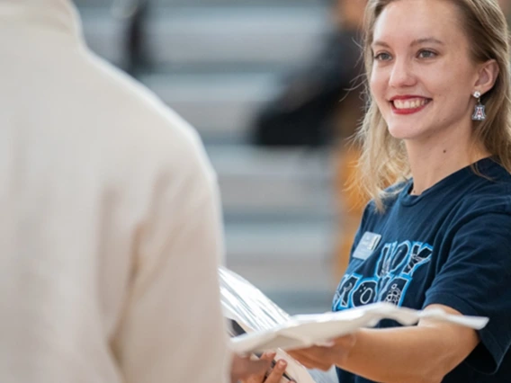 Smiling woman with long blonde hair wearing a navy blue t-shirt and University of Arizona earrings, handing papers to a person in a white sweater in a gymnasium setting. Let me know if you'd like these revised for a specific platform (like web accessibility, social media, or marketing use).