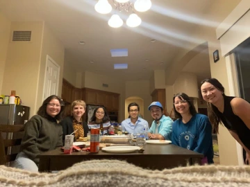 A group of eight people gathers around a dining table in a warmly lit home, smiling at the camera. The table is set with plates, drinks, and snacks, and the background features a kitchen with wooden cabinets. The atmosphere suggests a casual and friendly gathering.